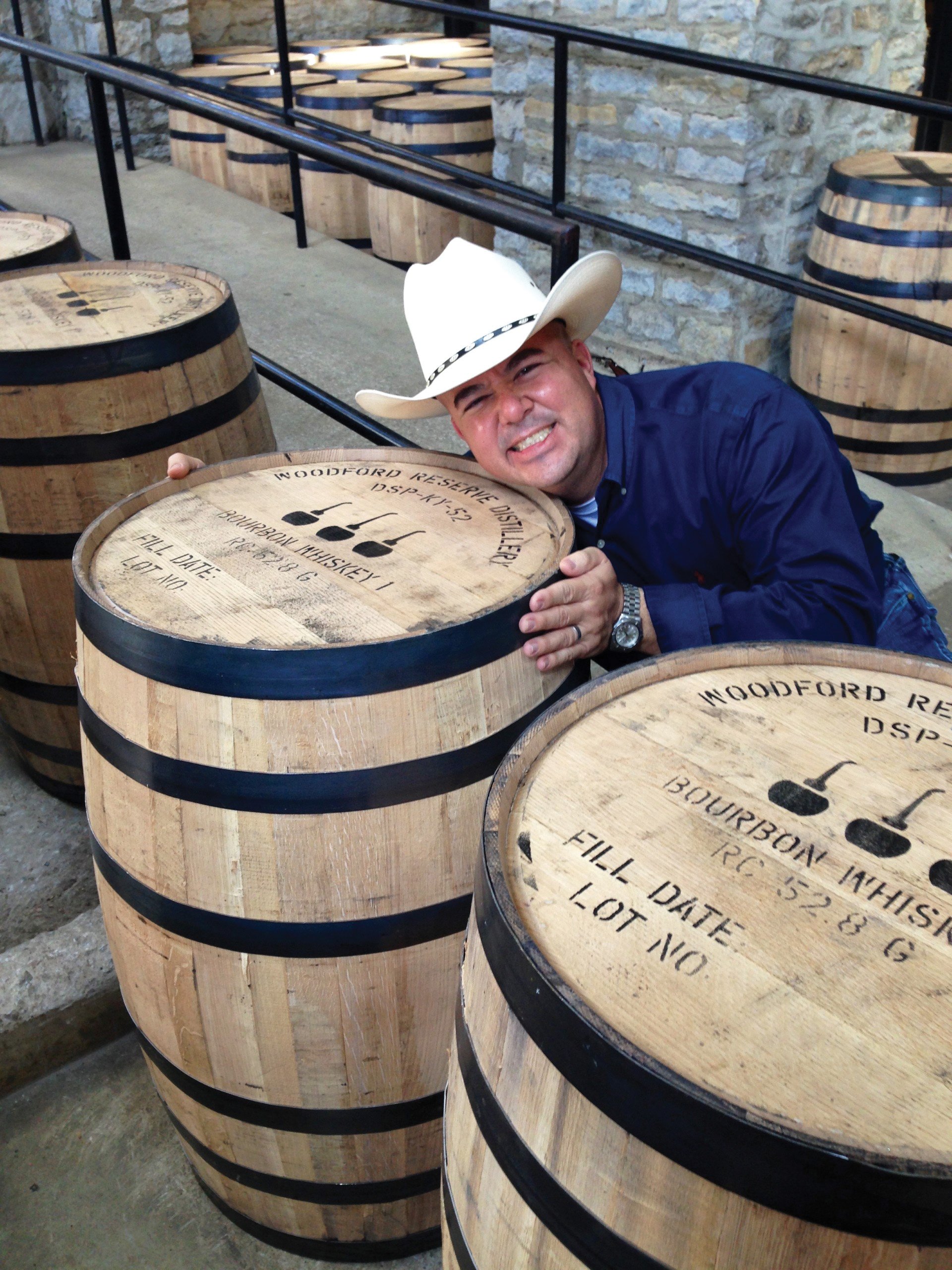 A man wearing a cowboy hat and a blue shirt is leaning over a large wooden barrel, smiling at the camera, in a facility with multiple barrels and stone walls.