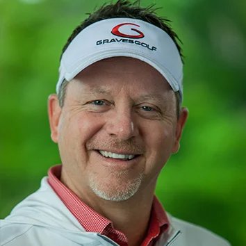 A smiling man wearing a white baseball cap with the Graves Golf logo, a red collared shirt, and a light gray jacket, outdoors with green trees in the background.