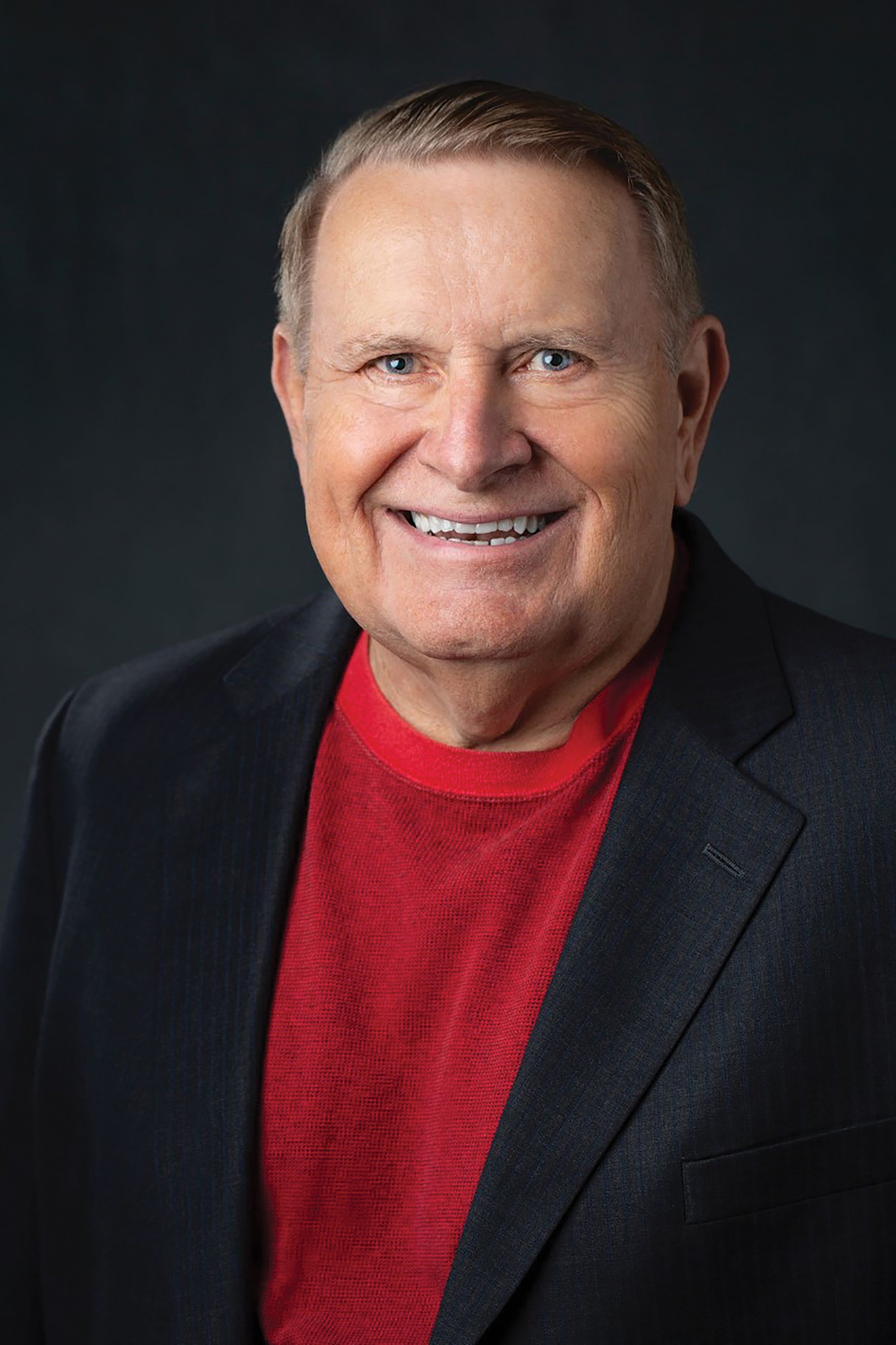 A smiling middle-aged man with blue eyes and short, light brown hair, wearing a dark blazer over a red shirt, set against a dark background.